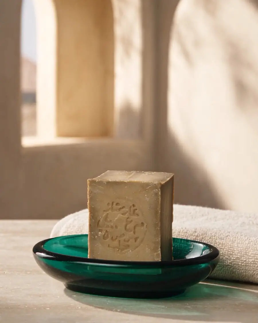 A bar of Aleppo soap on an emerald glass dish beside a folded towel